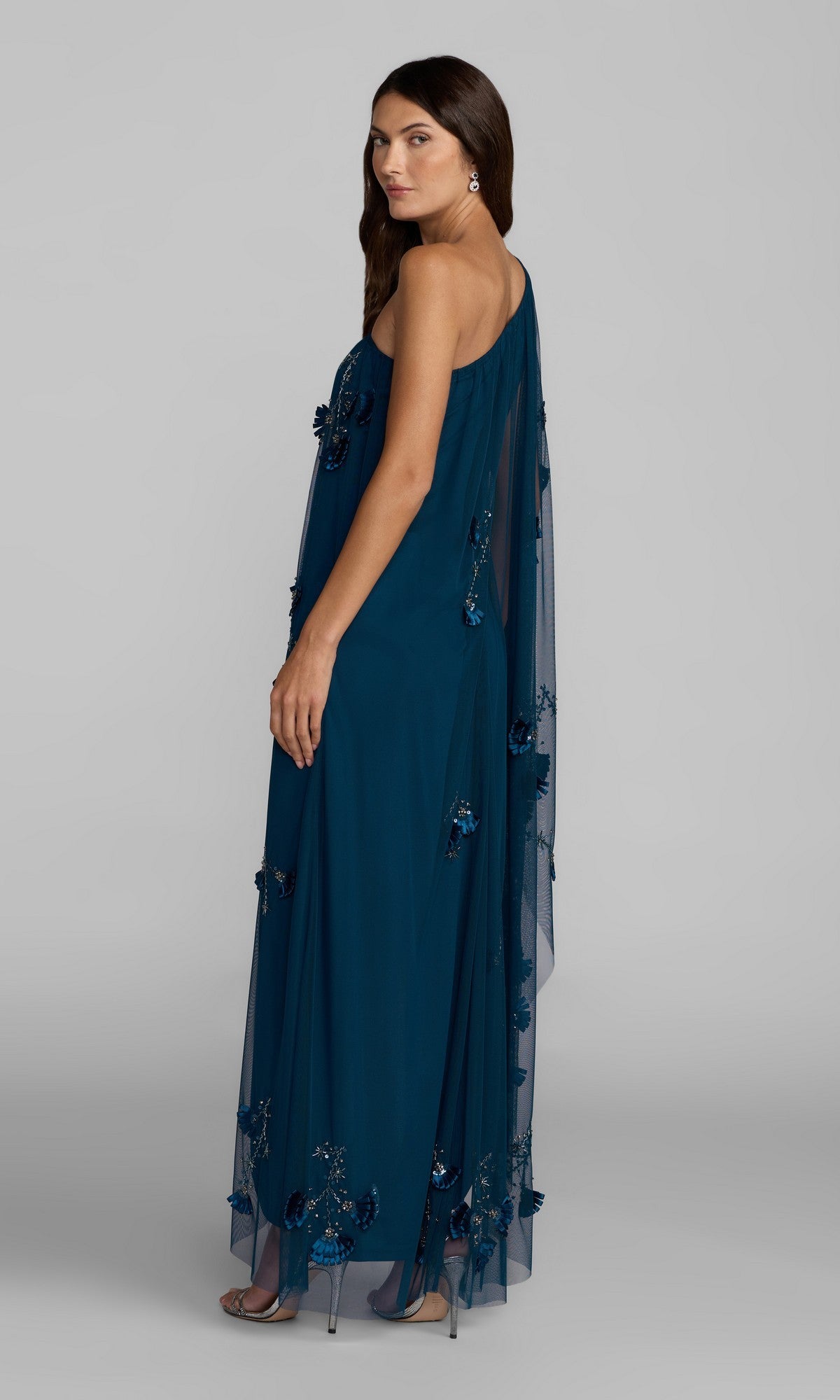 A woman, standing sideways against a plain background, wears the Long Formal Dress: Mac Duggal 9374—an elegant teal, one-shoulder gown adorned with floral embellishments and featuring a sheer cape sleeve.
